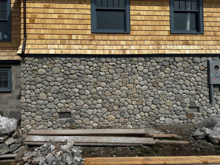 A house exterior with a stone wall base made of round gray stones, topped by tan wooden shingles. There are two large blue-trimmed windows and an electric utility box. Construction materials and debris are on the ground in front.