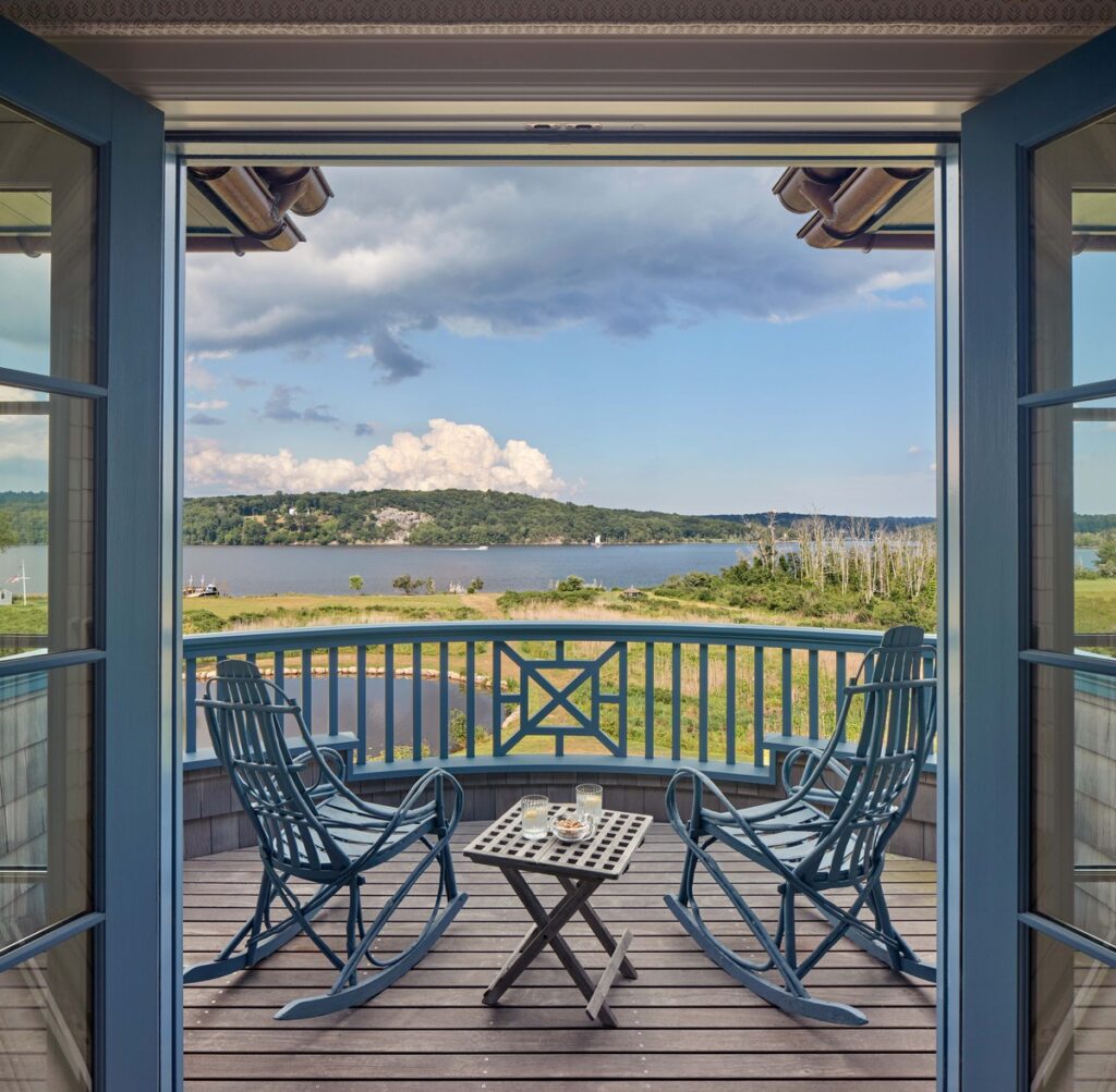 View from an open doorway onto a small balcony, part of inviting outdoor spaces, with two blue rocking chairs and a table with drinks and snacks. Beyond the railing is a scenic landscape of a river or lake, green hills, and a cloudy blue sky.
