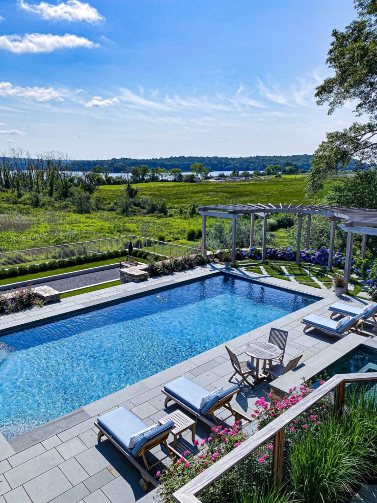 A bright, sunny day overlooks a rectangular swimming pool surrounded by lounge chairs and a patio with potted flowers, showcasing thoughtful outdoor design. Beyond the pool area, green fields and a river stretch toward trees and blue sky.