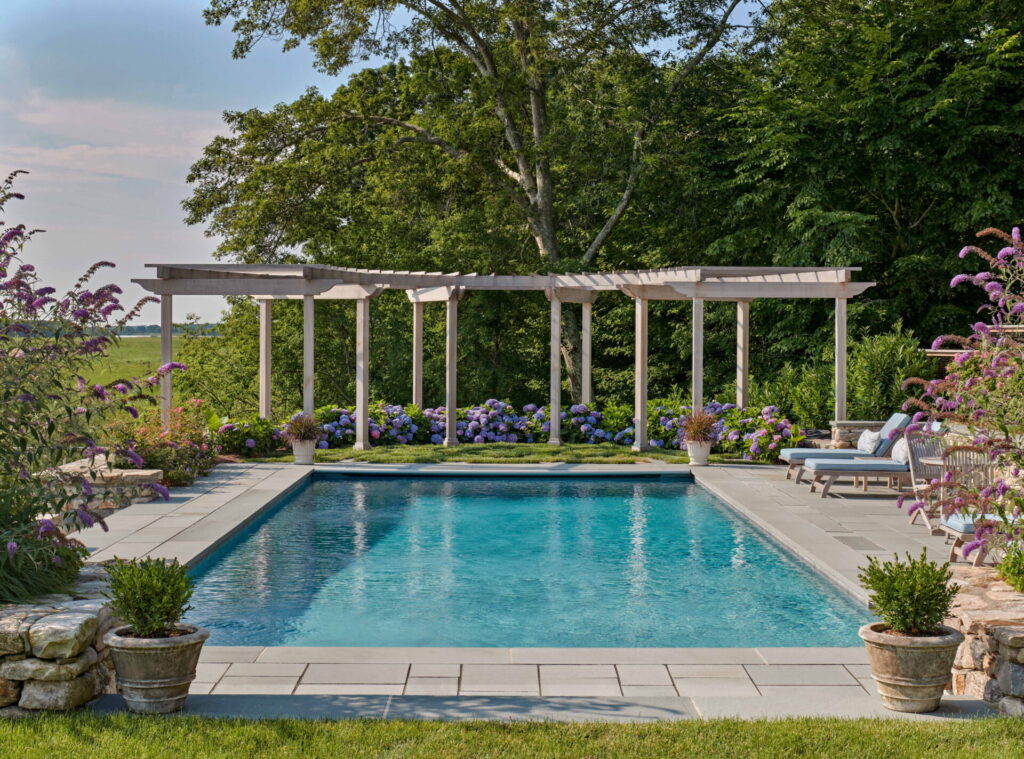A rectangular swimming pool is surrounded by a stone patio, potted plants, and blooming hydrangeas, creating inviting outdoor spaces. Lounge chairs and a pergola with climbing plants sit at the far end, backed by lush green trees and clear sky.