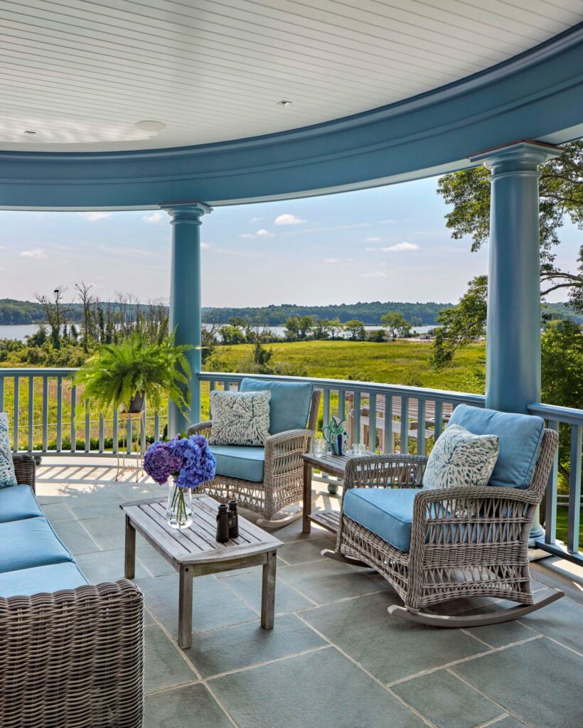 A covered porch with blue columns overlooks a scenic lake and greenery—one of our favorite outdoor spaces. Wicker sofas and chairs with blue cushions surround a wooden coffee table topped with purple flowers, while sunlight bathes the inviting, serene area.