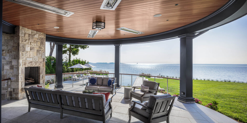 A covered patio with a wooden ceiling, stone fireplace, and modern outdoor furniture showcases thoughtful outdoor design. Glass railing offers an unobstructed view of the blue water and distant horizon, making it one of the favorite outdoor spaces.