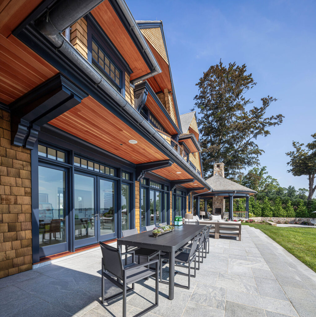 A modern patio showcases thoughtful outdoor design with a long black dining table and chairs beside a house with large glass doors, wood-paneled ceiling, and shingle siding. A stone gazebo with a fireplace elevates these favorite outdoor spaces amidst lawn and trees.