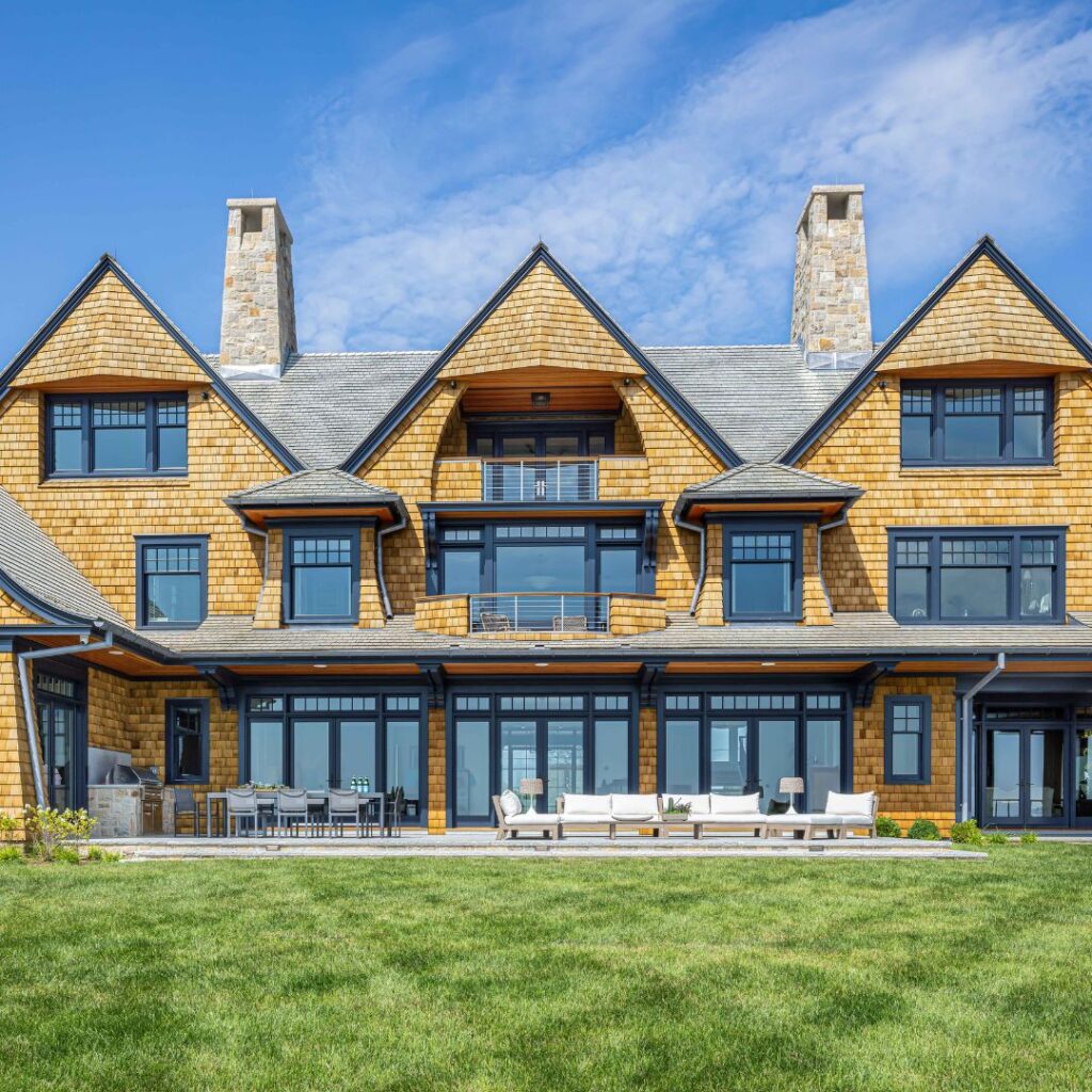 Large, three-story house with tan shingle siding, dark trim, and two stone chimneys. The facade features many large windows and balconies, with stylish outdoor decor on the patio seating and dining area, creating favorite outdoor spaces under a blue sky.