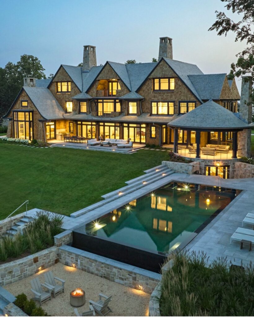 Large, elegant stone house with multiple gables and chimneys, glowing windows at dusk, and thoughtfully curated outdoor design. The landscaped backyard features a modern pool, hot tub, fire pit, and manicured lawn—one of our favorite outdoor spaces.