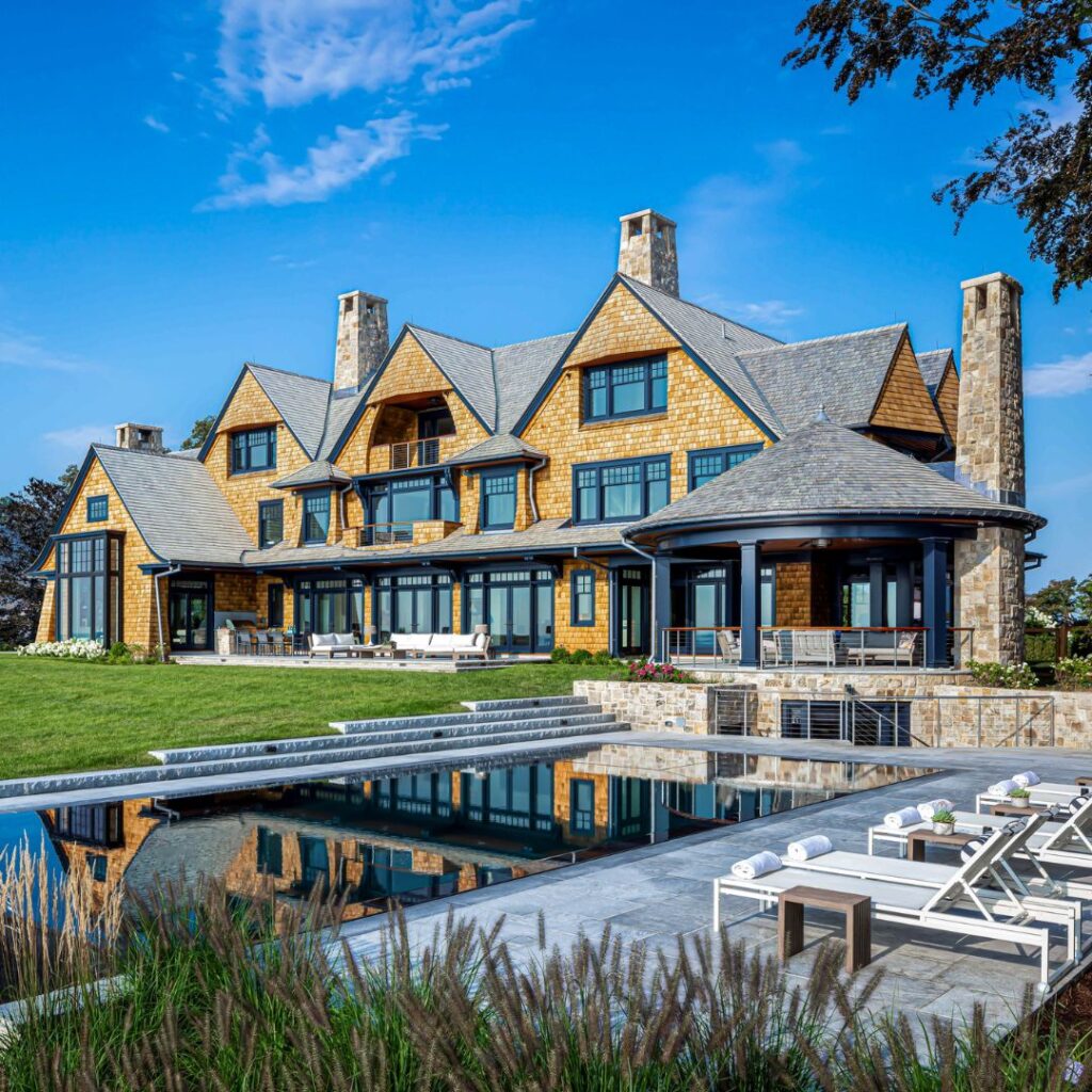 A large, elegant shingle-style house with stone chimneys, multiple gables, and large windows overlooks a rectangular swimming pool. The inviting outdoor spaces feature white lounge chairs with towels, lush lawn, and landscaped plants under a blue sky.