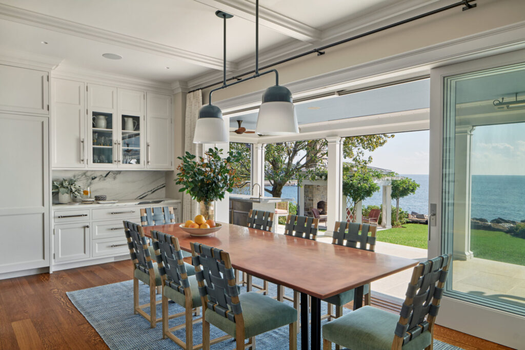 A modern dining room with a long wooden table, ten cushioned chairs, and two large pendant lights flows seamlessly into favorite outdoor spaces, as sliding glass doors reveal a patio with ocean views, green lawn, and blue sky.
