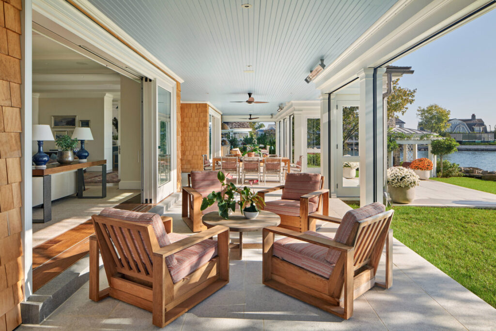 A spacious, sunlit patio highlights thoughtful outdoor design with four wooden armchairs around a round table. Sliding glass doors open inside, while outside features green grass, potted flowers, and a waterfront view—perfect for favorite outdoor spaces.