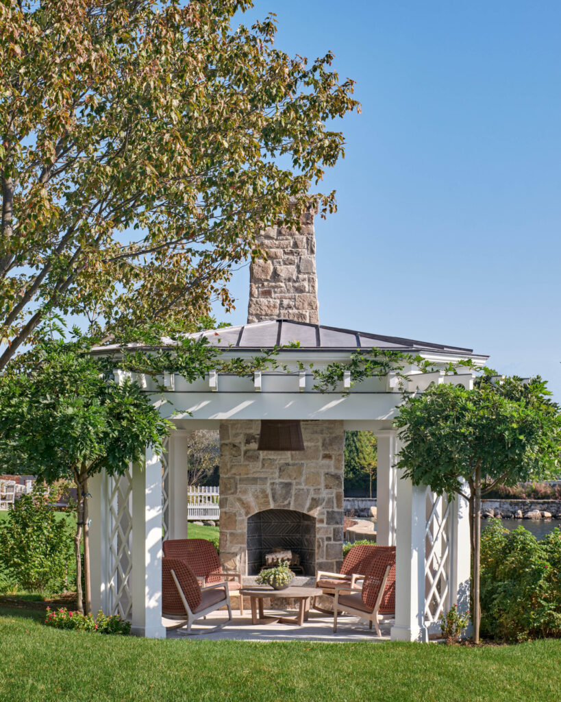 A white pergola with a tall stone fireplace, four red wicker armchairs, and a round wooden table creates one of our favorite outdoor spaces—an inviting outdoor design set on green grass, framed by trees, with water views under a clear blue sky.