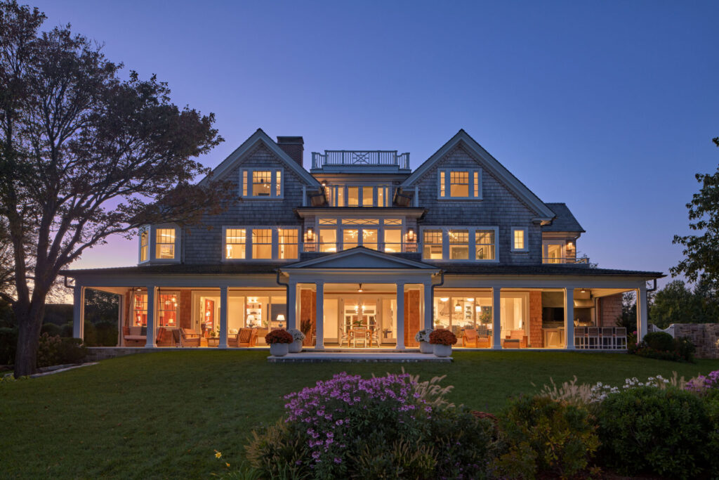 A large, elegant three-story house with many lit windows stands at dusk. The gray-shingled home features a wraparound porch and manicured lawn, showcasing thoughtful outdoor design among flower beds and trees beneath a clear sky.