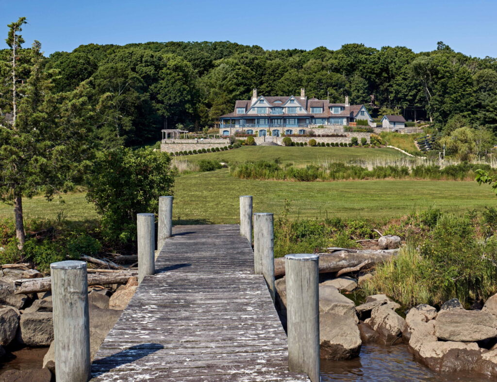 A weathered wooden dock leads over rocks and water to a stately house with a gray-blue roof, surrounded by lush green lawns and gardens—an inspiring example of outdoor design nestled under a clear blue sky.