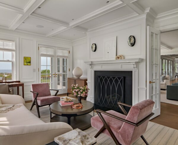 A bright, elegant living room with a coffered ceiling, pale walls, and wood floors. Pink velvet chairs and a cream sofa surround a black fireplace. A round coffee table holds flowers and decor; large windows reveal an ocean view. Art and clocks adorn the mantle.