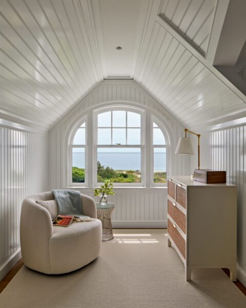 A cozy white reading nook with a curved armchair, side table, and wooden dresser sits under an arched window, offering a view of green foliage and the ocean beyond. Sunlight brightens the white paneled walls and ceiling, creating an inviting, peaceful space.