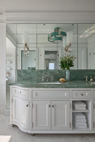 A white wooden bathroom vanity features a green marble countertop with an integrated sink and chrome fixtures. A large paneled mirror hangs above the counter, reflecting a teal drum chandelier and a glass vase holding a green plant. Below, open shelving holds neatly folded white towels.