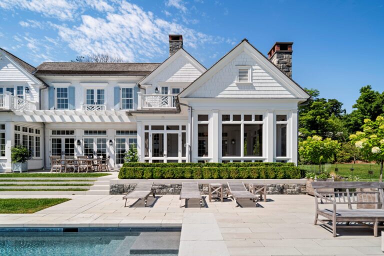 A white, multi-gabled house with blue shutters overlooks a stone patio and a swimming pool. The landscaped yard features trimmed hedges, three wooden lounge chairs, and a dining set on the patio, all set against a bright blue sky with wispy clouds.