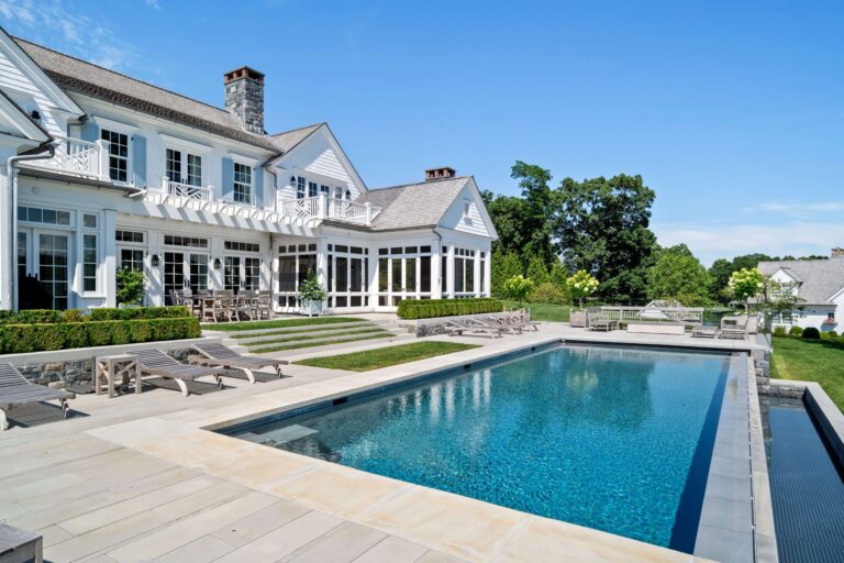 A large white house with a stone chimney overlooks a rectangular swimming pool set into a stone patio. Several wooden lounge chairs are arranged near the water, and tiered grassy steps lead up to the homes multiple French doors. The scene is set under a clear blue sky.