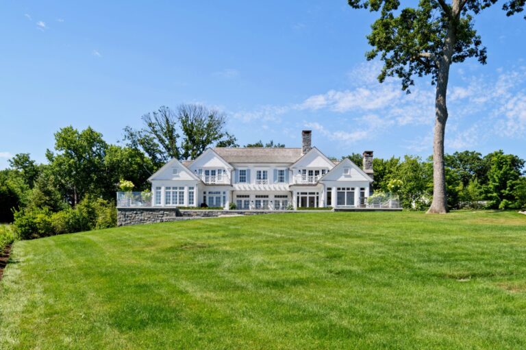 A large, white multi-story house sits behind a vast, expansive green lawn under a clear blue sky. The home features white siding, shuttered windows, balconies, and two stone chimneys, bordered by lush green trees on all sides.