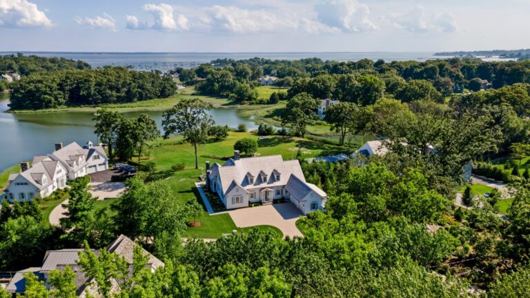 An aerial view shows a white, multi-gabled house with a light-colored roof centered on a lush green lawn. The property sits near a calm blue bay dotted with small boats, surrounded by dense trees and other large residential estates under a clear, sunny sky.
