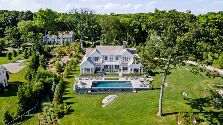 An aerial view of a large white two-story house with a grey roof, featuring a backyard pool and expansive stone patio. The residence is set within a lush green lawn surrounded by mature trees, with a prominent stone boulder sitting on the grass in the foreground.
