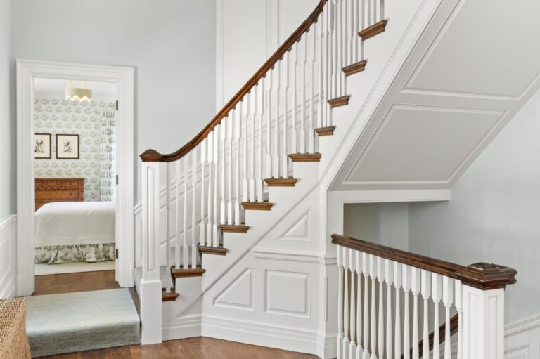 A white staircase with a stained wood handrail and matching treads ascends along a paneled wall in a brightly lit hallway. Through a doorway to the left, a bedroom is visible, featuring green patterned wallpaper, framed artwork, and a wooden headboard.