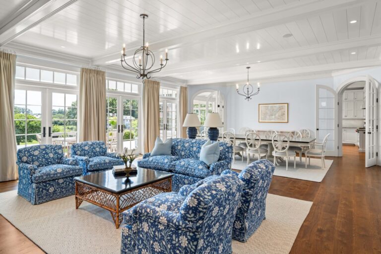 A bright living room features three armchairs and a sofa upholstered in blue and white floral fabric arranged around a dark wooden coffee table. In the background, a dining table with white chairs sits on a light rug, under white wood-paneled ceilings and iron chandeliers.