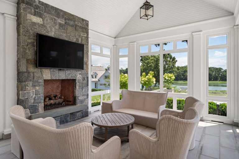 A bright sunroom features a stone fireplace with a mounted television and a seating area with light-colored wicker furniture. Large windows surround the space, offering a view of a lush green lawn, trees, and a lake in the distance.