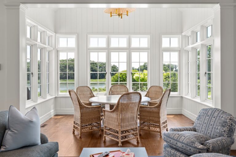 A sunroom features a round white table surrounded by four wicker chairs, centered beneath a gold lantern-style light fixture. Large windows line the walls, showcasing a green landscape and a pond. In the foreground, parts of two patterned blue armchairs frame the seating area.