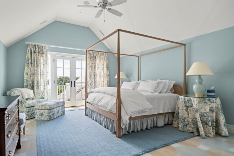 A bedroom features a light blue color scheme with a central wooden four-poster bed covered in white linens. A floral armchair and matching ottoman sit near glass doors, while a blue area rug covers the patterned floor. Two bedside tables with lamps flank the bed.