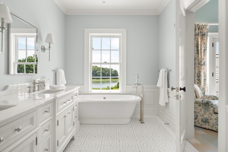 A bright bathroom features a white freestanding bathtub centered beneath a window overlooking a grassy lawn and trees. A long white vanity with a marble countertop sits to the left, while a doorway on the right reveals a cozy sitting area with floral furniture.