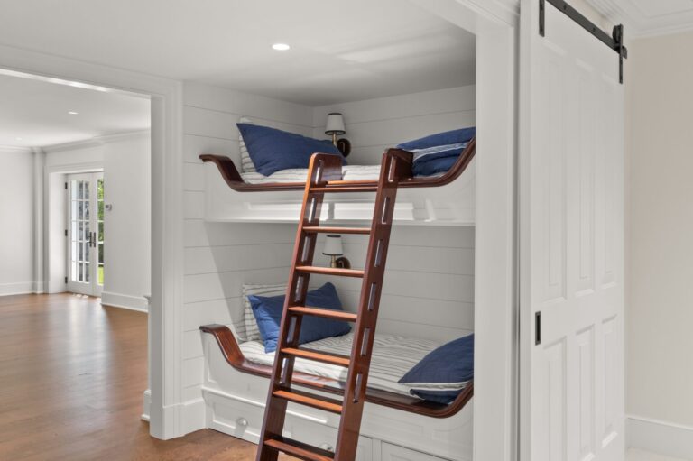 A built-in white bunk bed with dark wood trim is set into an alcove, featuring navy blue pillows and striped bedding. A dark wood ladder leans against the front, while a white sliding barn door is partially visible to the right.
