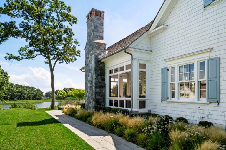 A white shingled house with light blue shutters features a tall stone chimney rising above a sunroom. A stone path curves alongside the foundation through ornamental grasses and flowers, overlooking a grassy lawn and a tranquil pond under a bright blue sky.