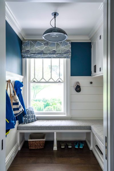 A mudroom nook features an L-shaped built-in bench with grey cushions beneath a large window. The walls are painted deep blue and white shiplap, accented by a geometric roman shade, a metal pendant light, and hooks holding bags, jackets, and a baseball cap.