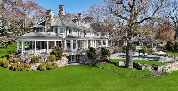A large, multi-story estate with gray siding and stone accents sits behind a vast, well-manicured lawn. To the right, a swimming pool and patio area are nestled near a mature tree and a secondary gazebo-style structure.
