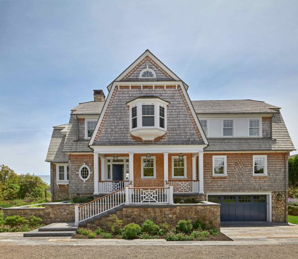 A large shingle-style house features cedar shake siding and a prominent central gable with a bay window. The home is elevated on a stone foundation with a front porch, a staircase with wooden railings, and a blue garage door, all set against a clear blue sky.