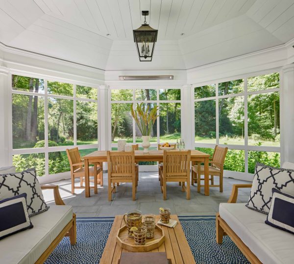 A sunroom features a wooden dining set surrounded by floor-to-ceiling windows overlooking a lush, green forest. In the foreground, two armchairs with white cushions and patterned navy pillows flank a low wooden coffee table set with a pitcher and glasses on a blue patterned rug.